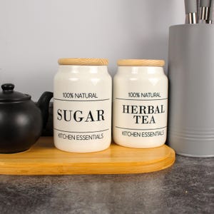 Op de afbeelding: Twee crèmekleurige keramische keukenpotten met houten deksels, met de labels "SUGAR" en "HERBAL TEA". De potten maken deel uit van de "KITCHEN ESSENTIALS" collectie en staan op een houten dienblad, met een zwarte theepot en een grijze keukengerei houder in de buurt.