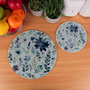 May include: Two round decorative glass trivets with a floral design. The trivets feature a light blue background with blue and white flowers and green leaves. One trivet is larger than the other. The trivets are on a wooden surface.