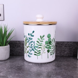 May include: A white cylindrical metal canister featuring a botanical print of various green eucalyptus and fern leaves. The container has a flat bamboo lid topped with a small, round, white knob handle, providing a secure cover for the storage vessel.