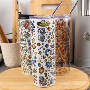 May include: Three insulated tumblers with vibrant, quirky designs. One has a space theme with rockets and planets. Another has a popcorn and movie theme. The third has a music theme. Each tumbler has a stainless steel straw.