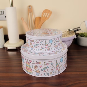Peut inclure: Deux boîtes à gâteaux rondes en métal blanc empilées. Elles sont décorées d'un motif de fête d'anniversaire coloré avec des gâteaux, des ballons et des confettis. Les boîtes sont posées sur une surface en bois.