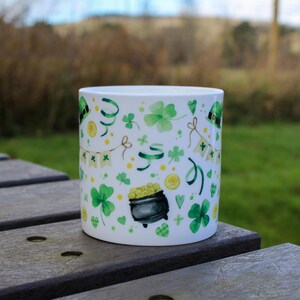 Op de afbeelding: Een witte keramische mok met een patroon van groene klaverbladeren en gouden munten. De mok staat op een houten tafel.