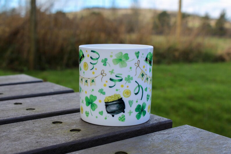 Op de afbeelding: Een witte keramische mok met een patroon van groene klaverbladeren en gouden munten. De mok staat op een houten tafel.