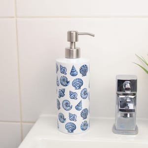 Può includere: Dispenser di sapone in ceramica bianca con pompa in acciaio inossidabile. Il dispenser è decorato con illustrazioni di conchiglie blu. C'è un rubinetto cromato accanto.