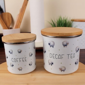 May include: Two white metal canisters with wooden lids. One says "COFFEE" and the other says "DECAF TEA" in black text. Both are decorated with sheep and green dots.