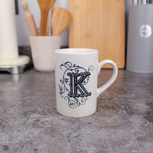 Puede incluir: Taza de cerámica blanca con una letra "K" negra y gris en un diseño ornamental. La taza tiene un asa curvada. El fondo muestra utensilios de cocina y una tabla de cortar.
