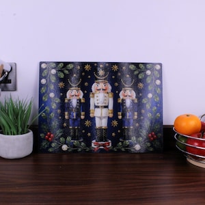 May include: A rectangular glass chopping board featuring a dark blue background with three nutcracker figures. The central nutcracker wears a white uniform, while the two flanking figures wear blue uniforms. Green holly leaves, red berries, and gold stars decorate the border.