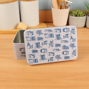 Könnte beinhalten: Eine rechteckige, hellgraue Metallbox mit Klappdeckel. Deckel und Seiten sind mit blauen Illustrationen von Vintage-Kameras verziert. Die Box ist geöffnet und gibt ihr Inneres frei. Im Hintergrund befinden sich Küchenutensilien.