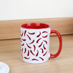 Può includere: Tazza in ceramica bianca con bordo e manico rossi. La tazza presenta un motivo ripetuto di peperoncini rossi su sfondo bianco.