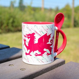 Puede incluir: Una taza de cerámica blanca con interior y asa rojos, con un diseño de dragón rojo. La taza incluye una cuchara roja a juego. El exterior tiene un patrón de salpicaduras verdes y rojas. La taza está sobre una superficie de madera.