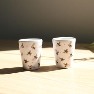 Puede incluir: Dos vasos de cerámica blancos con un patrón repetido de abejas ilustradas. Las abejas son negras y amarillas con alas grises. También hay pequeños puntos naranjas en los vasos.