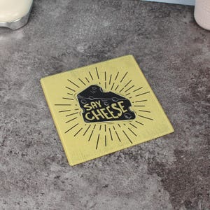 Può includere: Sottobicchiere quadrato giallo con un disegno a forma di spicchio di formaggio nero e la scritta "SAY CHEESE" in grassetto. La grafica del formaggio è circondata da linee radiali, creando un effetto a raggiera. Il sottobicchiere è probabilmente in vetro o materiale simile.