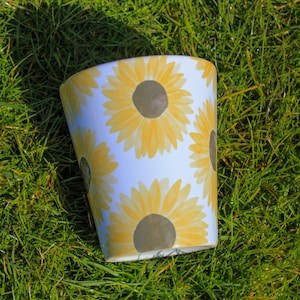 Può includere: Vaso in ceramica bianca con un motivo a girasole. Il vaso ha girasoli gialli e marroni su uno sfondo bianco.