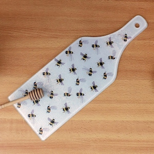 Può includere: Un tagliere da portata in vetro bianco con un design a forma di ape. Il tagliere ha la forma di una bottiglia di vino e ha un manico. Le api sono nere e gialle e sono sparse su tutto il tagliere.
