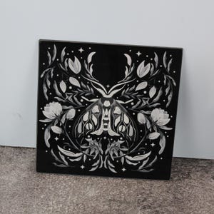 May include: Square black decorative art piece with a white moth illustration and floral details. The symmetrical design has a glossy finish. The artwork is suitable for wall decor and measures approximately 30 cm x 30 cm.