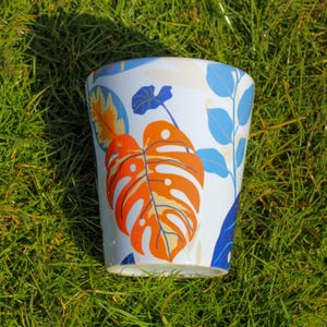 May include: A white ceramic planter with a tropical leaf design. The planter features blue, orange, and yellow leaves on a white background.