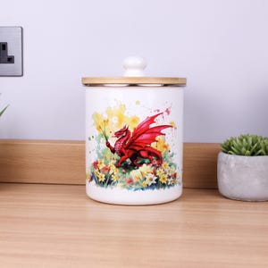 Puede incluir: Bote de cerámica blanco con tapa de bambú. El bote presenta una ilustración vibrante de un dragón rojo en medio de flores coloridas y salpicaduras de pintura de acuarela. El dragón sostiene una flor amarilla.