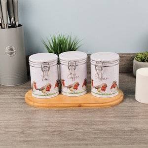 May include: Three white metal storage canisters with lids and clasps, labelled "Tea", "Coffee", and "Sugar". Each canister features a design of robins and sits on a wooden tray. A gray utensil holder is in the background.