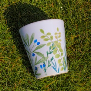 May include: White ceramic mug with a floral design featuring green leaves and blue flowers. The mug is sitting on a bed of green grass.