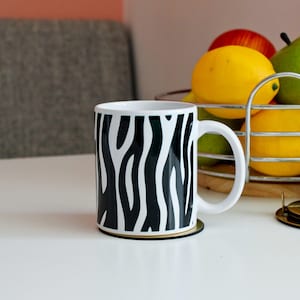 May include: A white ceramic mug featuring a bold black zebra stripe pattern wrapping around the exterior. The mug has a smooth cylindrical body and a curved handle on the right side, resting on a circular coaster on a white surface.