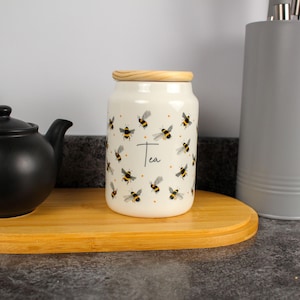 Op de afbeelding: Een witte keramische theepot met een houten deksel, versierd met zwart-gele bijenillustraties en het woord "Tea". De pot staat op een houten dienblad naast een zwarte theepot.