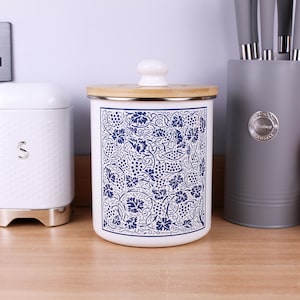 May include: A cylindrical white canister featuring a detailed blue pattern of grapevines and clusters of grapes. The container has a flat wooden lid with a small white knob handle centred on top. The design covers the front surface of the canister.