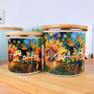 Peut inclure: Deux boîtes en céramique avec des couvercles en bambou. Les boîtes ont un motif de tournesol et de fleurs avec les mots "Coffee" et "Decaf" écrits dessus.