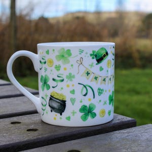 Op de afbeelding: Witte keramische mok met een groen, geel en wit ontwerp met klavertjesvier, gouden munten en een kabouterhoed. De mok staat op een houten tafel.