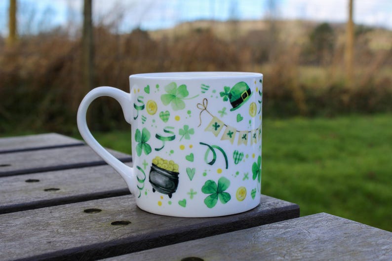 Op de afbeelding: Witte keramische mok met een groen, geel en wit ontwerp met klavertjesvier, gouden munten en een kabouterhoed. De mok staat op een houten tafel.