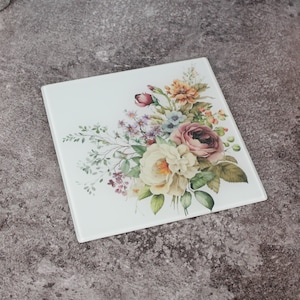 Può includere: Piastrella decorativa quadrata in vetro bianco con un motivo floreale. La composizione floreale comprende rose e altri fiori nei toni del rosa, giallo, arancione e viola, con fogliame verde. Adatta per l'uso come sottopentola o elemento decorativo. Misura circa 20 cm x 20 cm.