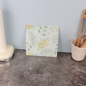 Op de afbeelding: Vierkante glazen onderzetter met een botanisch ontwerp. De onderzetter heeft een witte achtergrond met een patroon van groene bladeren en gele bloemaccenten. Het is ontworpen om oppervlakken tegen hitte te beschermen.