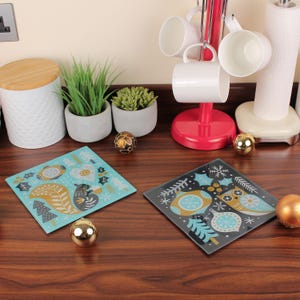 Könnte beinhalten: Zwei quadratische Glasuntersetzer mit festlichen Designs. Einer ist hellblau mit einem Eichhörnchen- und Baummuster. Der andere ist dunkelgrau mit einem Eulen- und Ornamentmuster. Goldene und bronzefarbene Dekorationskugeln sind ebenfalls vorhanden.
