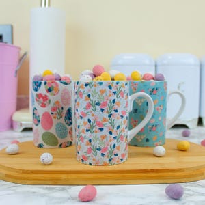 May include: Three ceramic mugs with different floral and egg patterns filled with pastel-colored chocolate eggs. The mugs are on a wooden cutting board with more chocolate eggs scattered around.