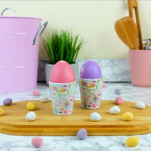 Peut inclure: Deux supports à œufs en céramique avec un motif floral et le texte "Spring Time" dessus. Les supports à œufs contiennent des œufs roses et violets. Plusieurs petits œufs en chocolat sont éparpillés sur une surface en bois.