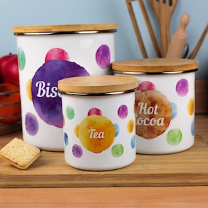 Puede incluir: Tres botes de cocina blancos con tapas de bambú. Cada bote presenta un diseño de acuarela con lunares de colores y etiquetas: "Bis", "Tea" y "Hot Cocoa". Los botes están sobre una superficie de madera.
