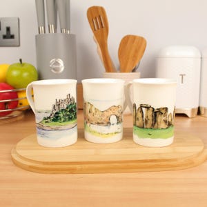 Peut inclure: Trois mugs en céramique blanche avec des illustrations de paysages. L'un représente un château, un autre une arche rocheuse et le troisième Stonehenge. Les mugs sont posés sur une planche de service en bois.