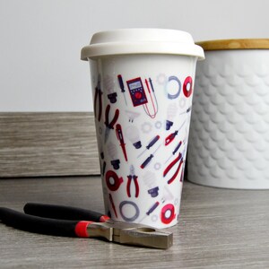 May include: White ceramic travel mug with a colorful design of tools and electrical components. The mug has a white lid.