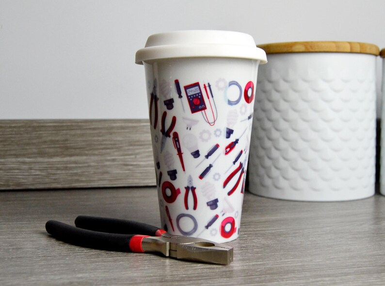 May include: White ceramic travel mug with a colorful design of tools and electrical components. The mug has a white lid.