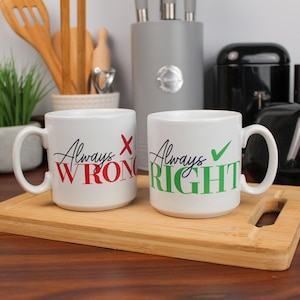 May include: Two white ceramic mugs with black handles. One mug says "Always Wrong" in red with a red X, and the other says "Always Right" in green with a green tick. The mugs sit on a wooden chopping board.
