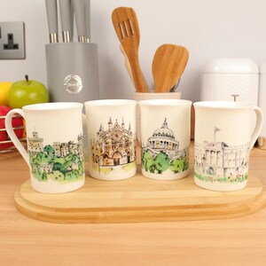 Peut inclure: Quatre mugs en céramique blanche, chacun orné d'une illustration architecturale différente. Les motifs comprennent un paysage urbain, une cathédrale, un bâtiment à dôme et un bâtiment gouvernemental. Les tasses sont disposées sur une planche à découper en bois.