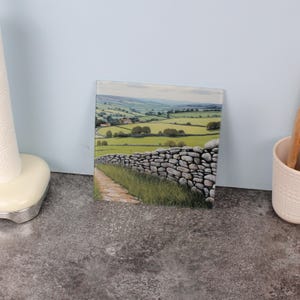 Puede incluir: Posavasos cuadrado de vidrio con pintura de paisaje. La obra representa un muro de piedra que bordea un camino a través de un campo verde, con colinas y cielo azul. Un artículo decorativo.