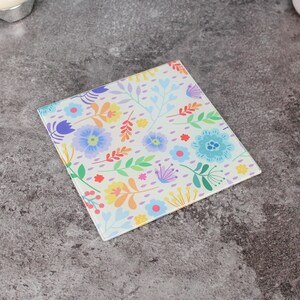 Può includere: Sottobicchiere quadrato in vetro con motivo floreale. Il design presenta fiori e foglie colorati nei toni del blu, viola, giallo e arancione su sfondo bianco. Un oggetto decorativo.