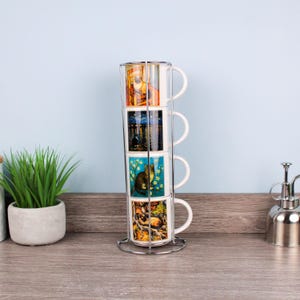 Può includere: Un set di quattro tazze impilate in ceramica bianca con diverse illustrazioni colorate, tenute in un supporto in metallo argentato. Le tazze presentano immagini di gatti, paesaggi e altri disegni decorativi.