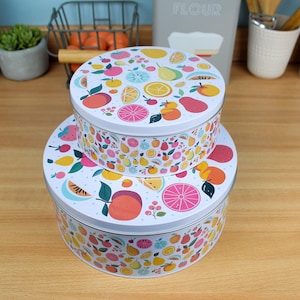 Peut inclure: Deux boîtes à gâteaux rondes en métal blanc empilées. Elles présentent un motif coloré de fruits variés, notamment des oranges, des fraises et des citrons. Ces boîtes sont conçues pour conserver les pâtisseries. Diamètres d'environ 20 cm et 25 cm.