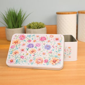 Puede incluir: Una caja de almacenamiento rectangular de metal blanco con un diseño floral con rosas rosas, naranjas y moradas y otras flores. La caja está abierta, revelando un interior liso. Hay un pequeño recipiente a juego al lado.