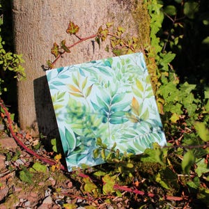 Op de afbeelding: Een vierkante glazen onderzetter met een aquarelontwerp van groene en gele bladeren. De onderzetter ligt op een bed van groen mos en bruine stenen.