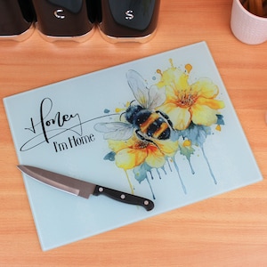 Könnte beinhalten: Ein blaues Schneidebrett aus Glas mit einem Aquarell-Design einer Biene auf gelben Blumen. Der Text "Honey I'm Home" ist in schwarzer Kursivschrift geschrieben.
