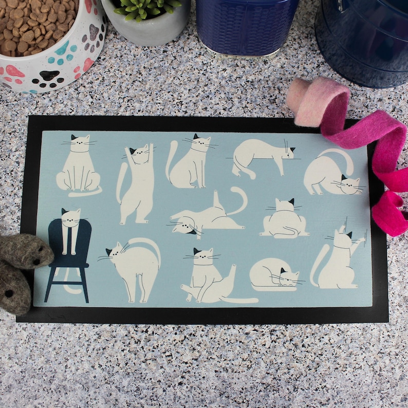 May include: A blue and white kitchen mat with a black border features a pattern of white cats in various yoga poses. The mat is on a light gray countertop.