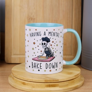 May include: A white ceramic mug with a light blue interior and handle. The exterior features a cartoon skeleton wearing a chef hat and apron, rolling dough on a pink board. The text reads HAVING A MENTAL BAKE DOWN, surrounded by small colourful stars.