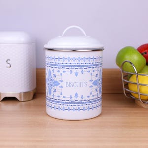 Peut inclure: Une boîte à biscuits cylindrique en métal blanc avec un couvercle et une poignée. La boîte présente un motif décoratif bleu et bleu clair avec des éléments floraux et géométriques. Le mot BISCUITS est imprimé en bleu sur le devant.